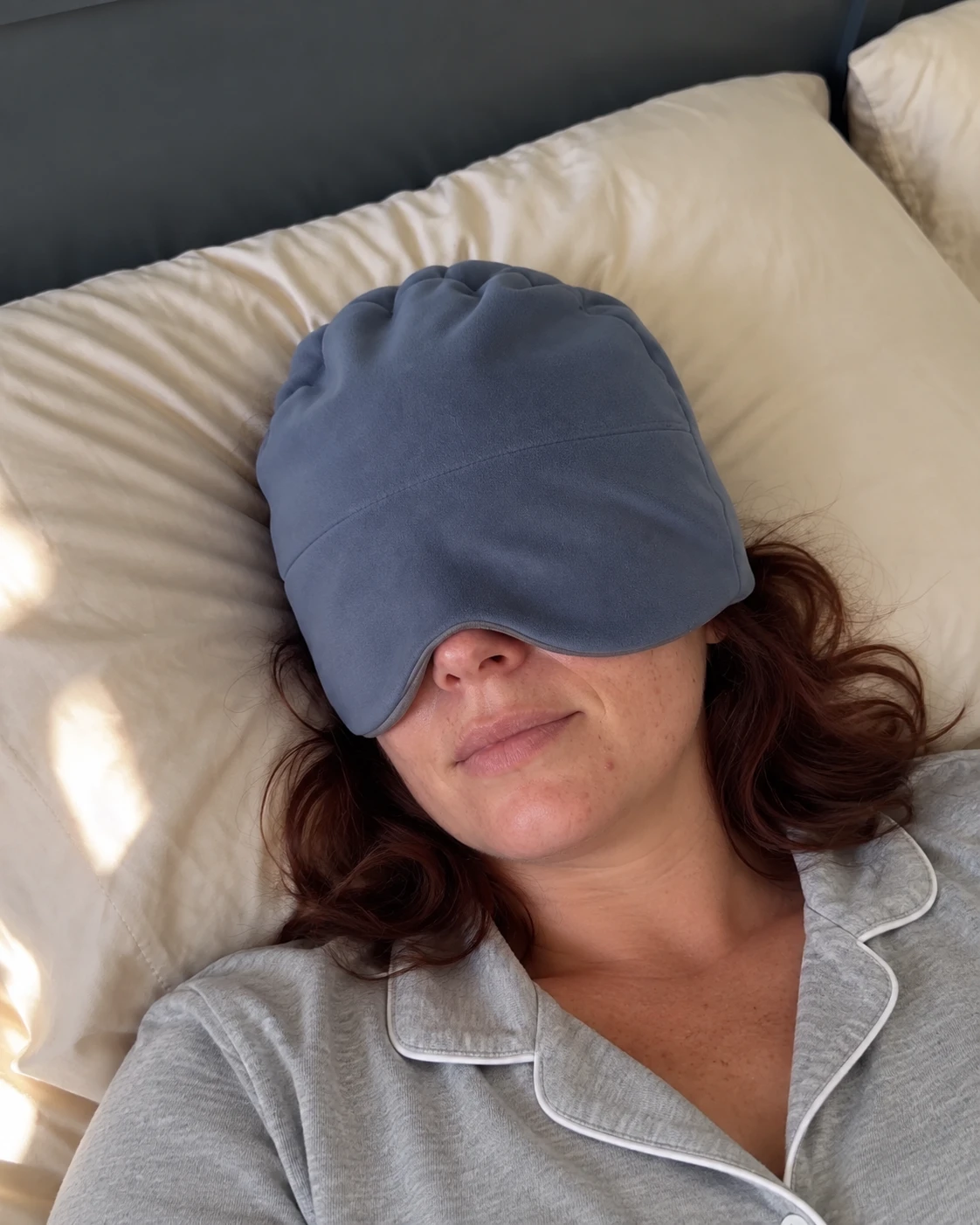 Woman resting on her back wearing the blue weighted sleep mask on a cream pillow