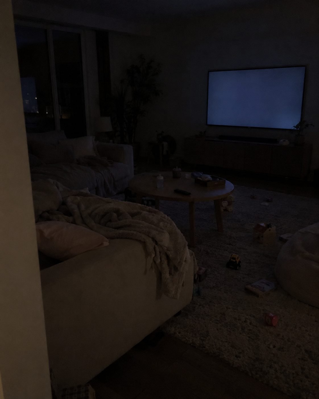 A dark living room late at night, lit only by an idle TV screen, toys on the rug